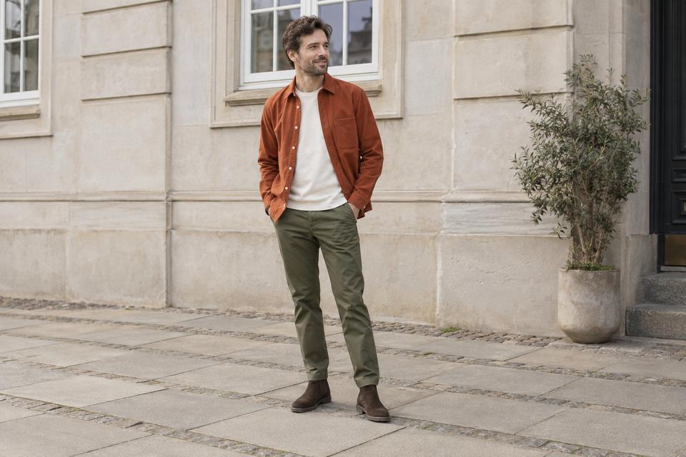 En mand med åben rustfarvet overshirt over råhvid T-shirt, olivengrønne chinos og mørkebrune ruskindsstøvler foran en lys facade.